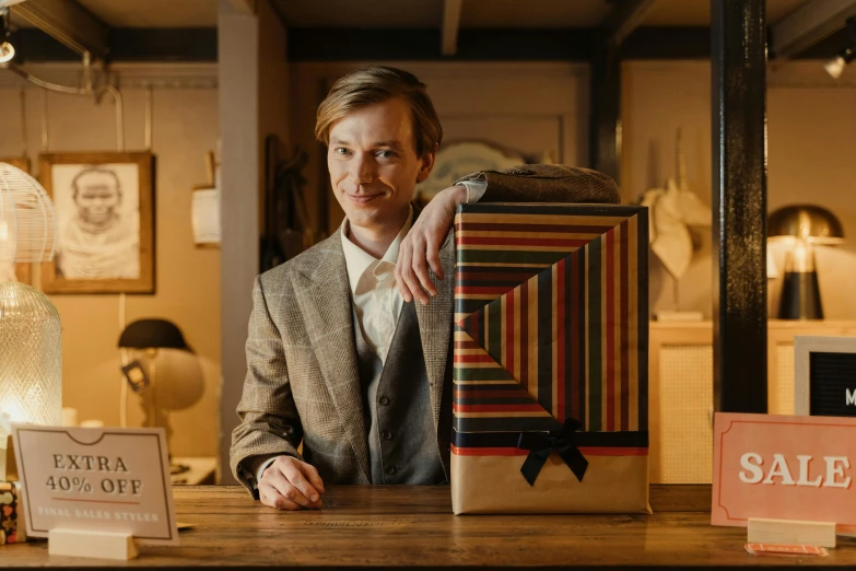a man sitting at a table with a box in front of him, by Alice Mason, pexels contest winner, wes anderson and gucci, wearing a pinstripe suit, holding gift, charlie cox