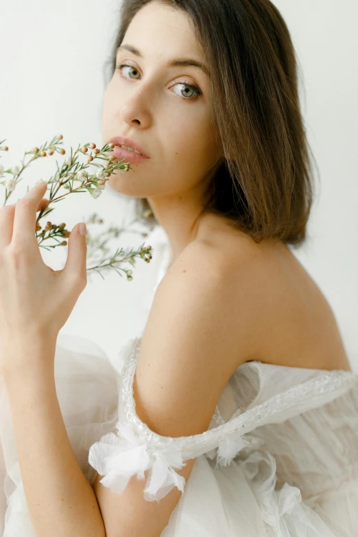 a woman in a white dress holding a flower, inspired by Elsa Bleda, trending on unsplash, white backdrop, natural lips, brunette, with soft bushes