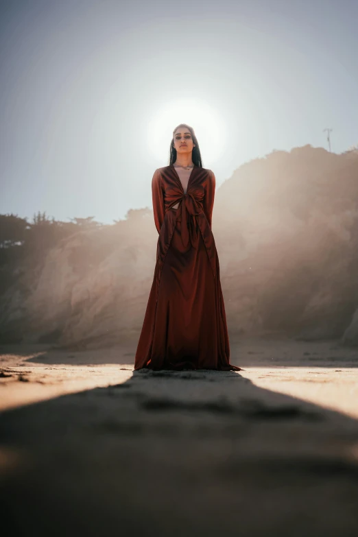 a woman in a long red dress standing in front of the sun, an album cover, pexels contest winner, standing on rocky ground, wearing brown robes, fashion shoot 8k, instagram post