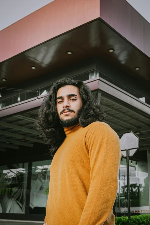 a man with long hair standing in front of a building, pexels contest winner, wearing turtleneck, wavy hair yellow theme, ash thorp khyzyl saleem, he is wearing a brown sweater