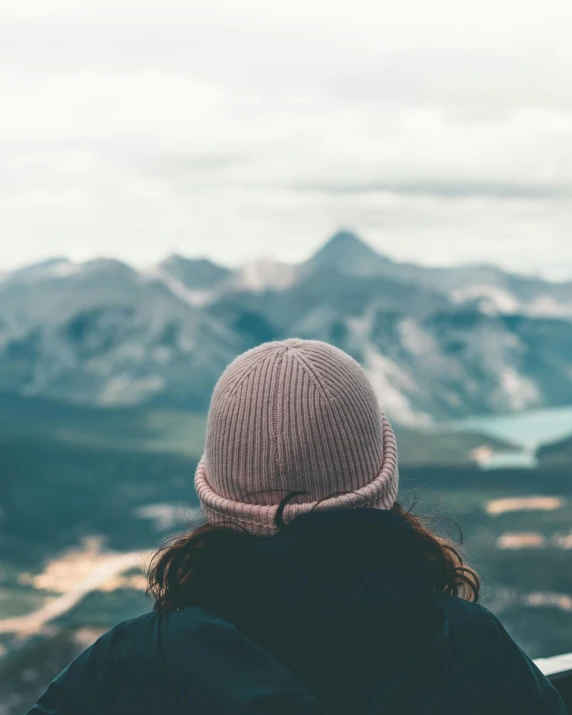 a woman standing on top of a mountain looking at a valley, a polaroid photo, by Emma Andijewska, trending on unsplash, beanie hat, curls on top of his head, in muted colours, banff national park