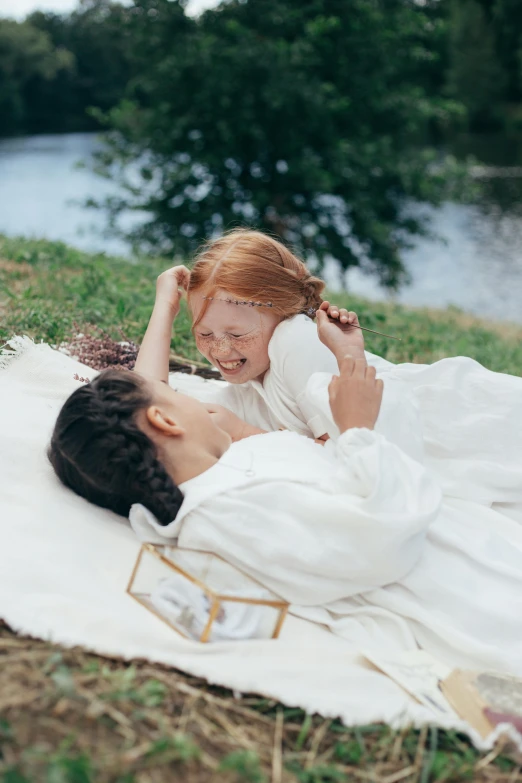 a woman laying on a blanket next to a little girl, inspired by Kate Greenaway, pexels contest winner, romanticism, wearing white robe, live-action archival footage, al fresco, smiling playfully