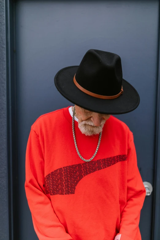 a man standing in front of a door wearing a hat, inspired by George Pirie, trending on pexels, modernism, long orange sweatshirt, red on black, willie nelson on stage, grandfatherly