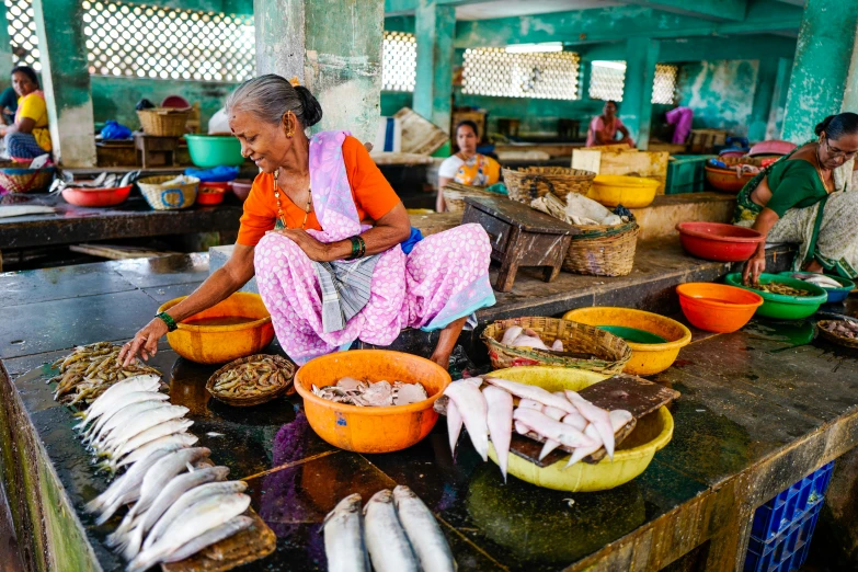 a woman sitting at a table with bowls of fish, by Julia Pishtar, pexels contest winner, sri lanka, fresh food market people, thumbnail, full body image