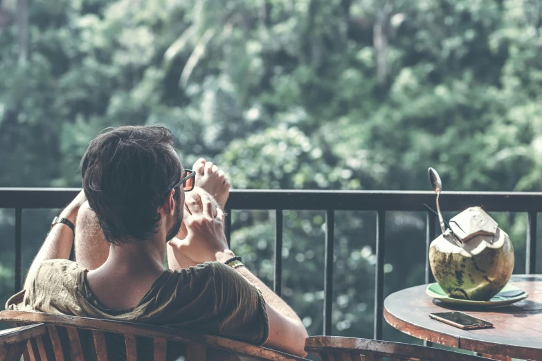 a man sitting on top of a wooden chair next to a table, pexels contest winner, in the background is lush jungle, balcony scene, taking a picture, spying discretly