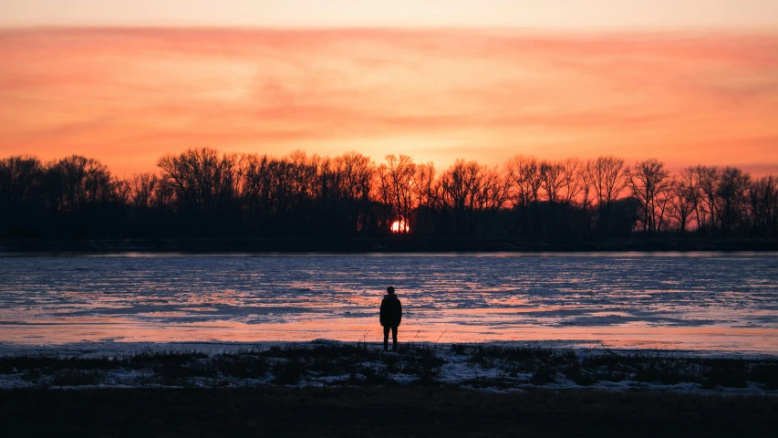 a person standing on a beach next to a body of water, by Attila Meszlenyi, pexels contest winner, cold sunset, silhouettes in field behind, sunset red and orange, river in front of him