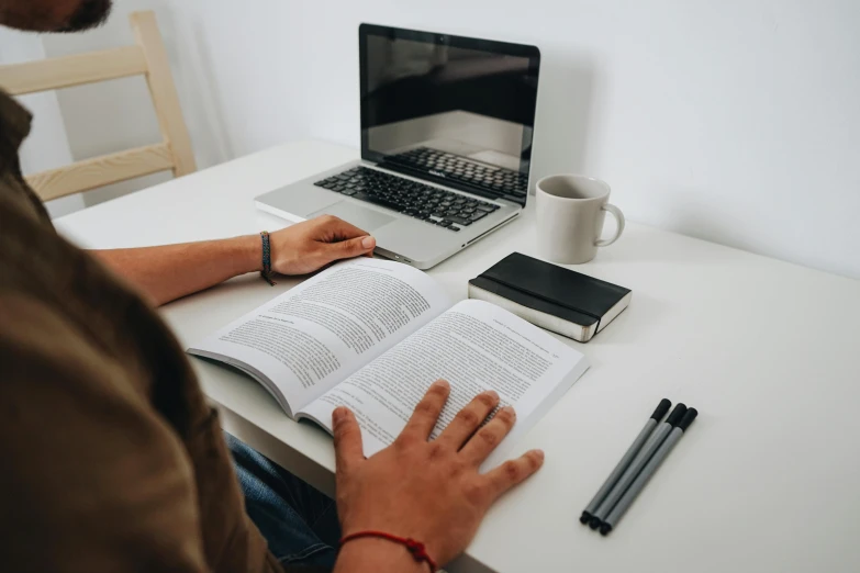 a man sitting at a table with a laptop and open book, pexels contest winner, textbooks, 🦑 design, background image, ground level shot