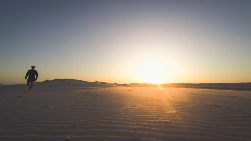 a man walking across a sandy beach at sunset, unsplash contest winner, white desert background, ultrawide angle, bright sun bleached ground, australian desert