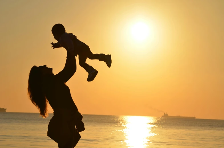 a woman holding a baby up in the air, pexels contest winner, symbolism, sunset at the beach, toys, thumbnail, no cropping