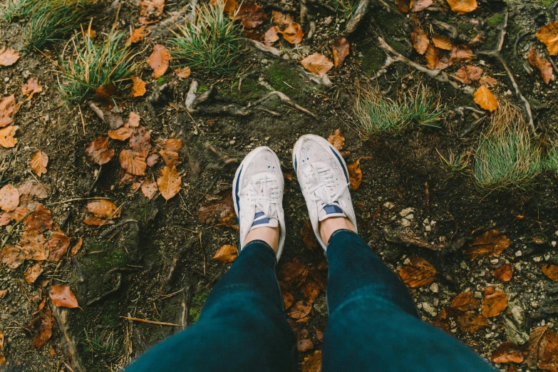 a person standing on top of a leaf covered ground, pexels contest winner, blue jeans and grey sneakers, 🍂 cute, in woods, white shoes