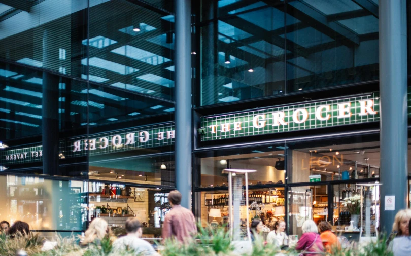 a group of people walking in front of a store, inspired by Edwin Georgi, unsplash, food court in a mall, large green glass windows, sitting down, te pae