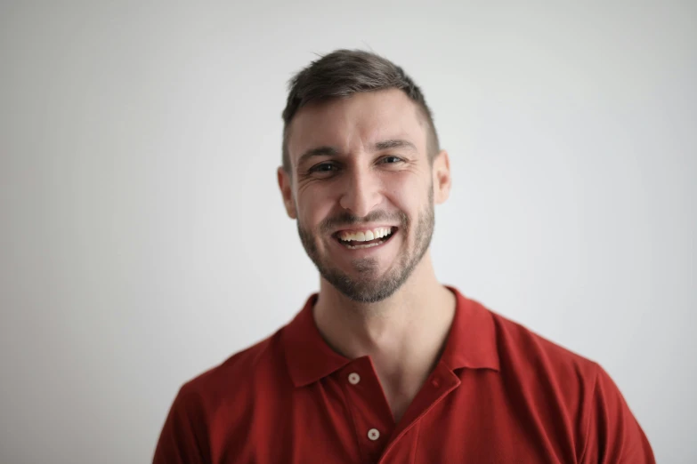 a man in a red shirt smiles at the camera, by Adam Marczyński, profile image, on a gray background, stjepan sejic, smiling at camera