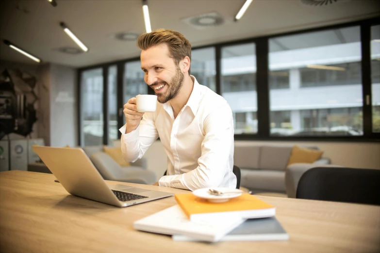 a man sitting at a table with a laptop and a cup of coffee, with an elegant smile, lachlan bailey, thumbnail, central hub