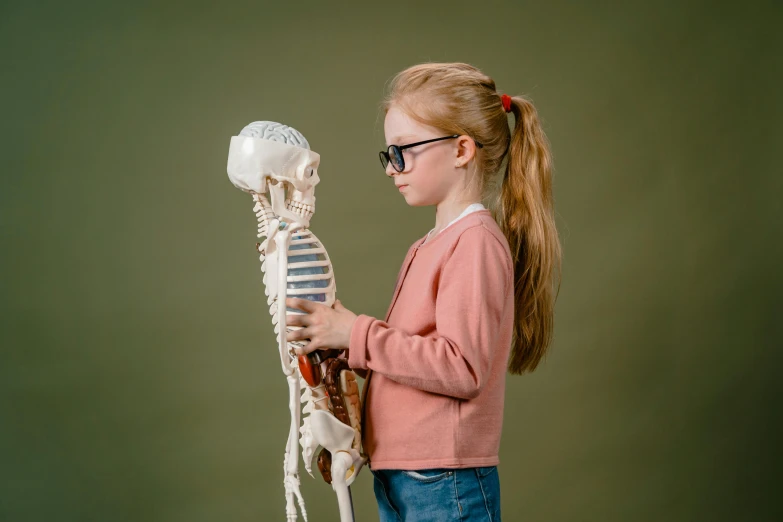 a little girl holding a model of a human skeleton, a picture, inspired by Sarah Lucas, shutterstock, glasses |, mary jane ansell, high quality picture, with square glasses