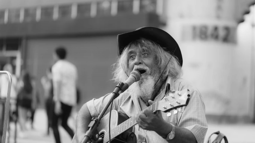 a man playing a guitar and singing into a microphone, a black and white photo, by Joze Ciuha, overalls and a white beard, in times square, long grey hair, aboriginal capirote