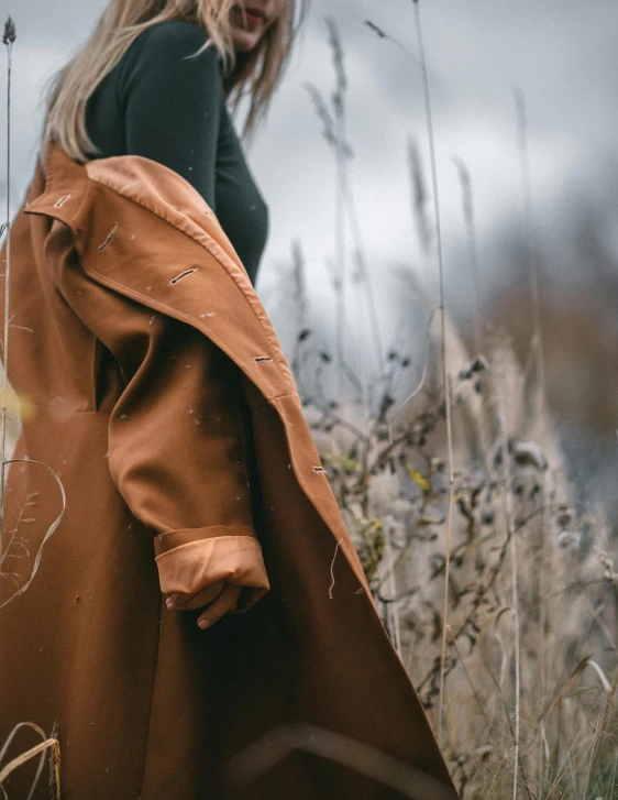 a woman standing in a field of tall grass, by Emma Andijewska, trending on unsplash, visual art, wearing a brown leather coat, wearing trenchcoat, detailed product shot, half image
