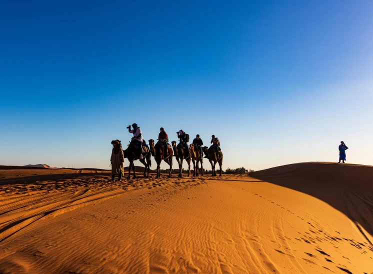 a group of people riding camels across a desert, by Julia Pishtar, pexels contest winner, hurufiyya, blue sky, australian, warm glow, group of seven