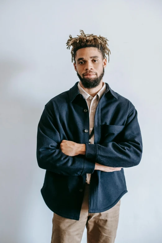 a man with dreadlocks standing in front of a white wall, an album cover, by Edward Bailey, trending on unsplash, in a navy blue sweater, he wears a big coat, button up shirt, zachary corzine
