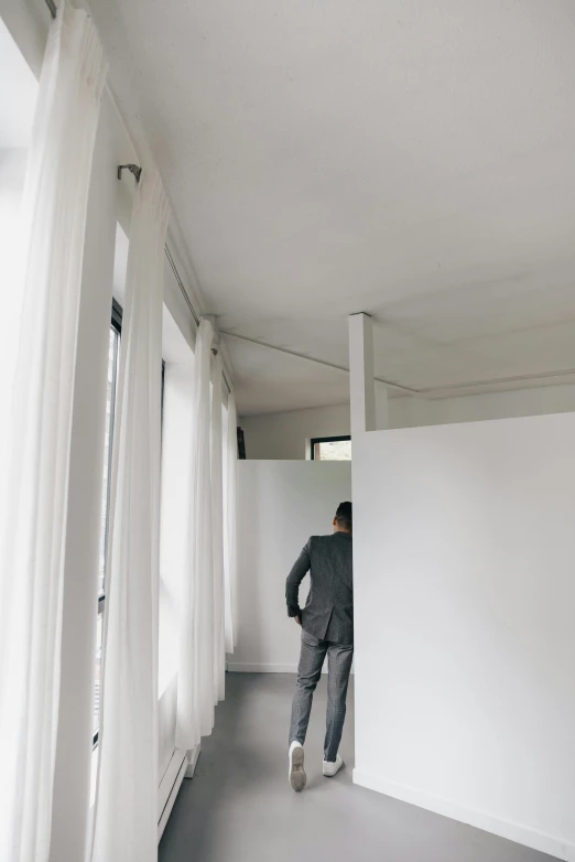 a man in a suit standing in an empty room, unsplash, modernism, curtains, white room, cubicles, hiding