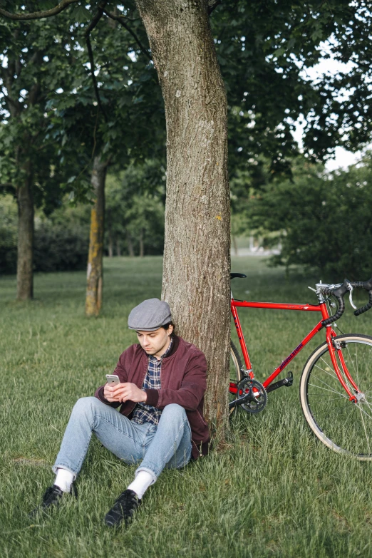 a man sitting under a tree next to a bike, looking at his phone, wearing newsboy cap, screenwriter, thoughtful )