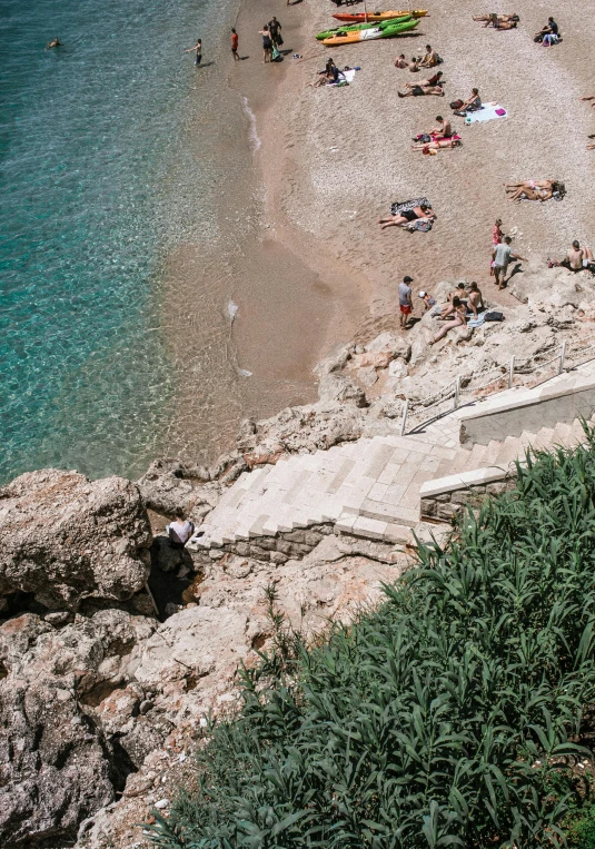 a group of people laying on top of a sandy beach, long winding stairs going down, boka, coastal cliffs, jen atkin