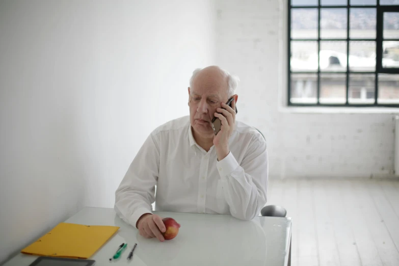 a man sitting at a table talking on a cell phone, in a white room, old man doing hard work, ian callum, with apple