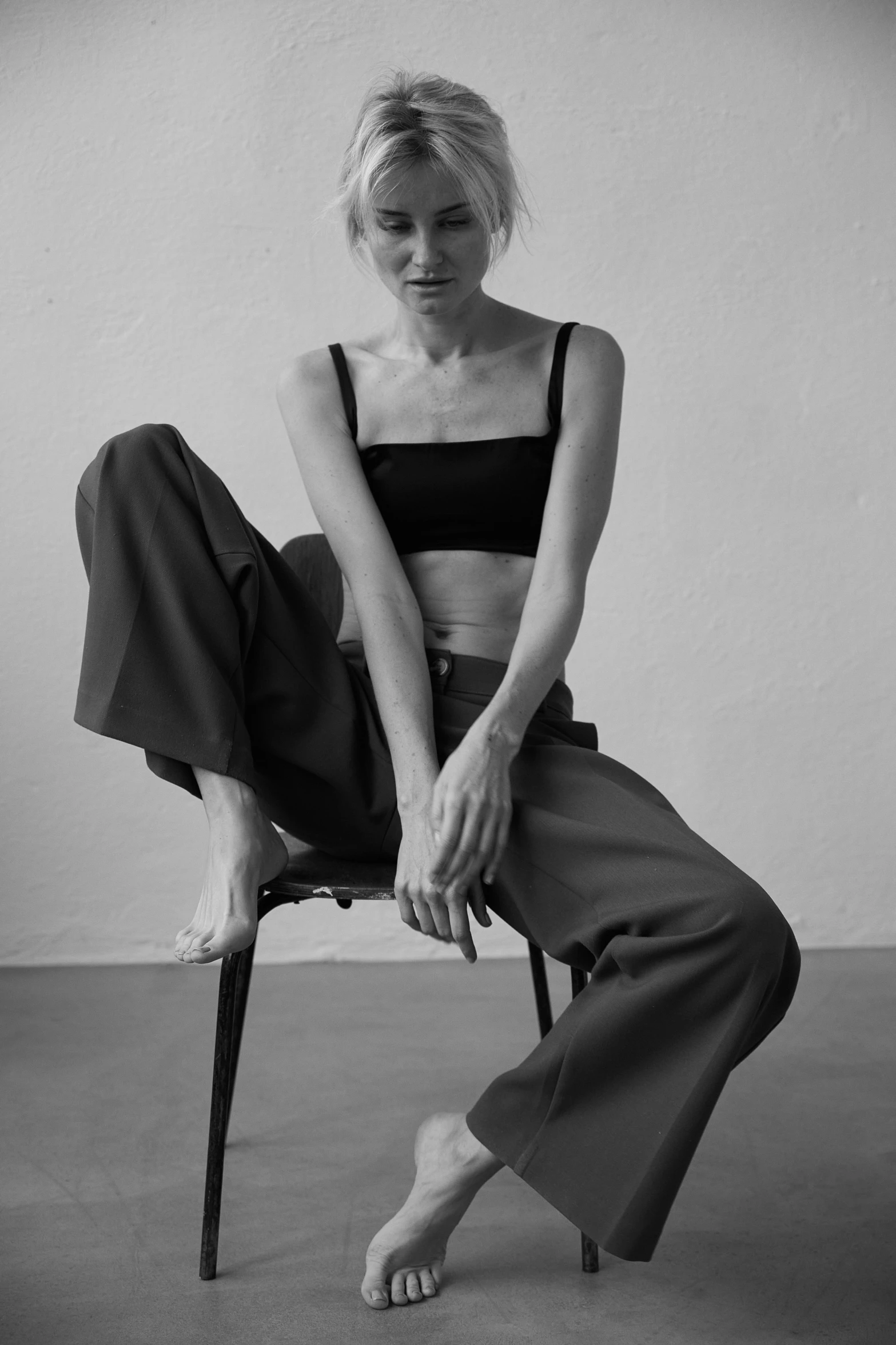 a black and white photo of a woman sitting on a chair, a black and white photo, unsplash, photorealism, black trousers, anorexic figure, wearing a cropped top, editorial photo from magazine