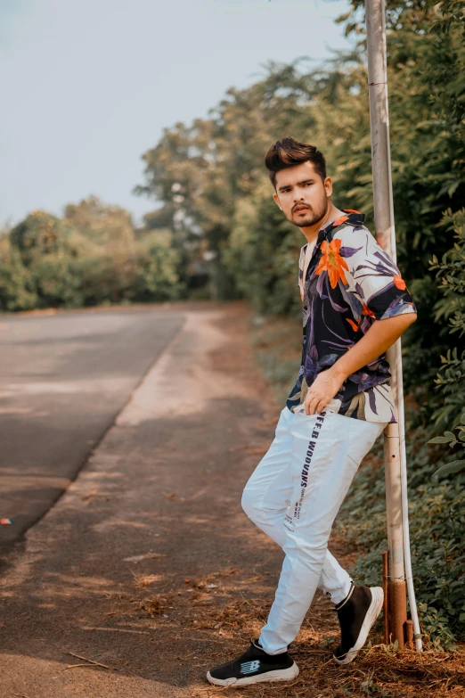 a man leaning against a pole on the side of the road, an album cover, by Adam Dario Keel, trending on pexels, patterned clothing, indian super model, tropics, wearing a shirt and a jean