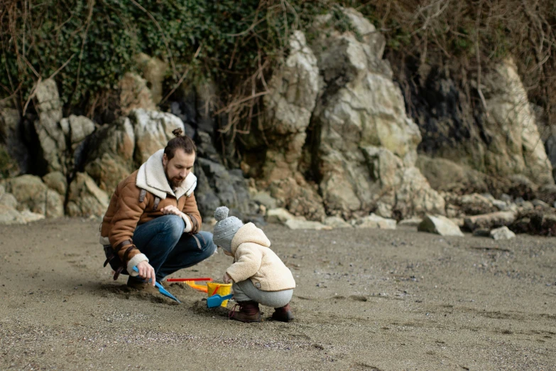 a man playing with a teddy bear on the beach, by Arabella Rankin, pexels contest winner, plein air, father with child, standing on rocky ground, grey, multicoloured