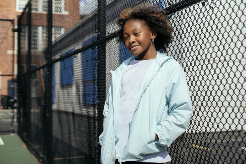 a girl standing in front of a fence on a tennis court, by Nina Hamnett, unsplash, blue parka, light skinned african young girl, wearing a light blue shirt, confident pose