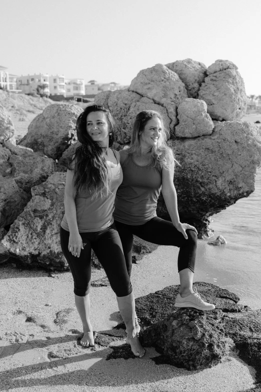 a couple of women standing next to each other on a beach, by Alexis Grimou, standing pose on stones, low quality photo, yoga, monochrome:-2