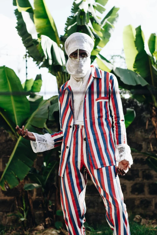 a man dressed in a red, white and blue striped suit, an album cover, inspired by Ras Akyem, afrofuturism, in style of juergen teller, wearing white pajamas, stripe over eye, official store photo
