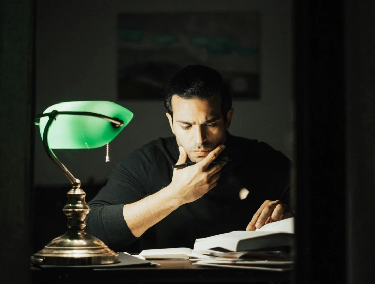 a man sitting at a desk reading a book, a colorized photo, inspired by Germán Londoño, pexels contest winner, glowing green lights, joe taslim, profile image, emerging from a lamp