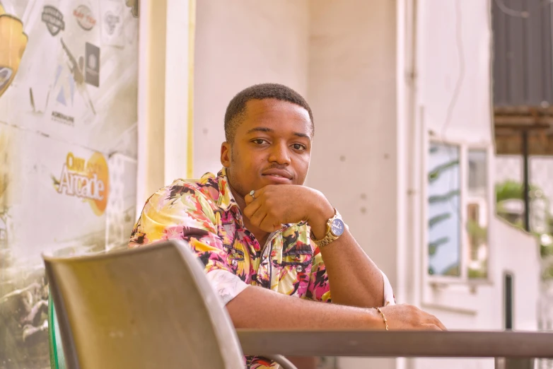 a man sitting at a table in front of a wall, wearing yellow floral blouse, emmanuel shiru, avatar image, high quality image