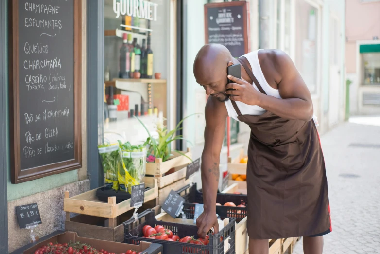 a man standing in front of a store talking on a cell phone, by Barthélemy Menn, pexels contest winner, wearing an apron, muscular bald man, permaculture, adut akech