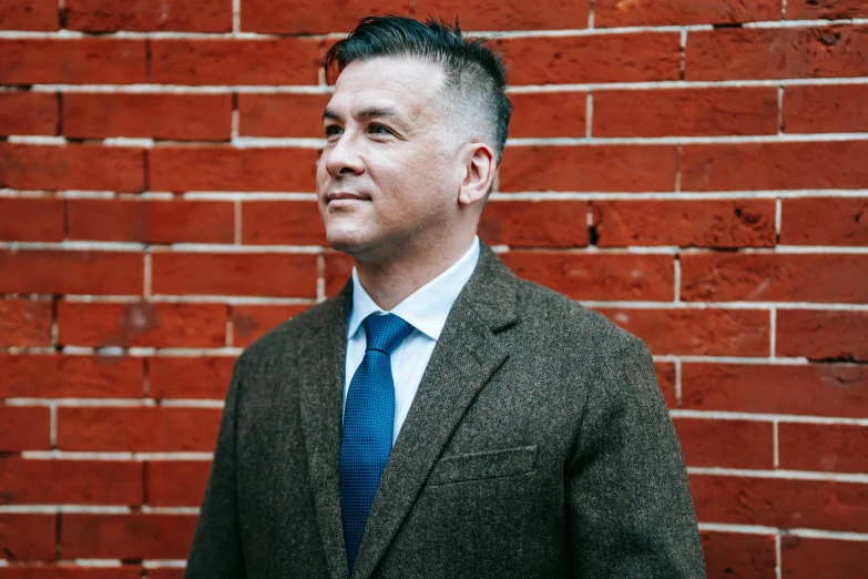 a man in a suit and tie standing in front of a brick wall, darren shaddick, mansik yang, profile image