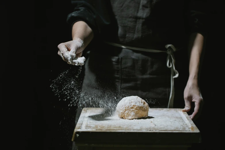 a person sprinkling powder on a doughnut on a cutting board, a portrait, unsplash, holding a baguette, white clay, dark and dim, looking across the shoulder