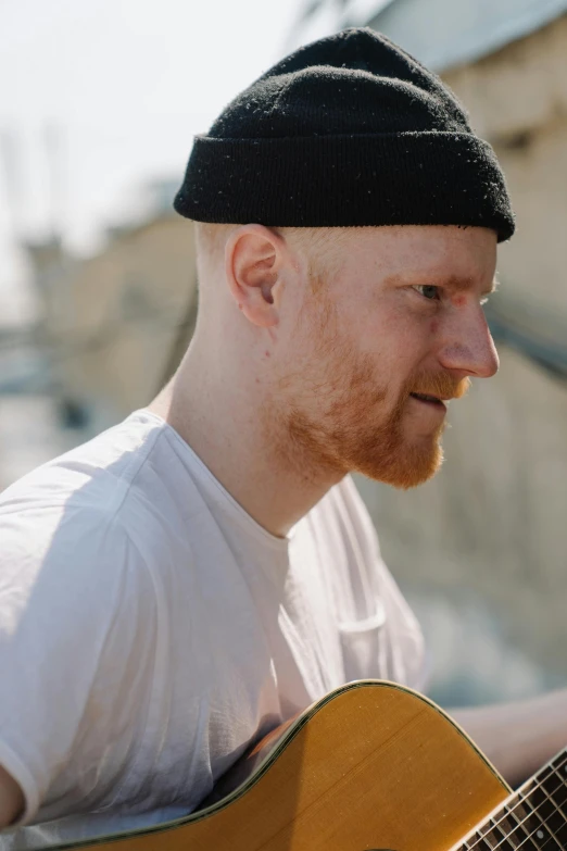 a man with a beard playing a guitar, ginger hair with freckles, bald on top, looking to the side off camera, slightly sunny