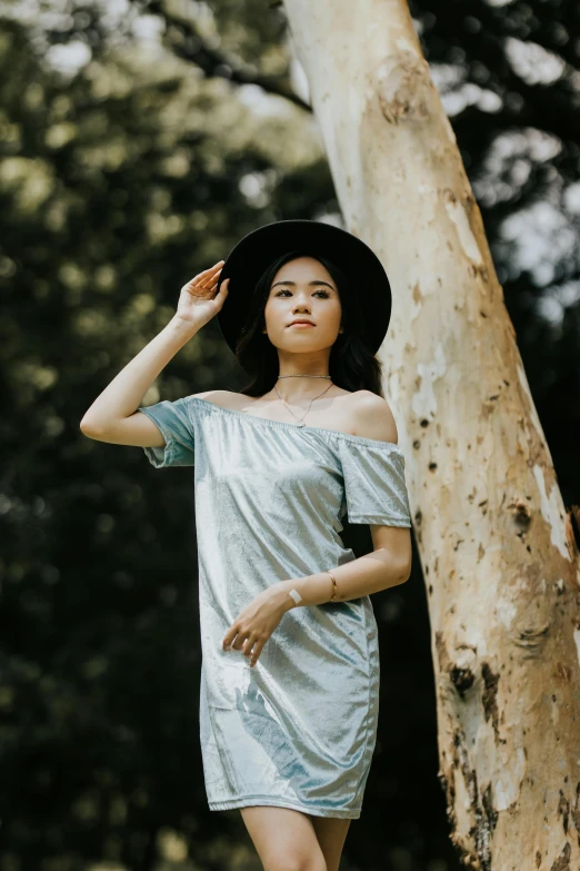 a woman standing next to a tree wearing a hat, inspired by helen huang, unsplash, wearing silver dress, wearing casual clothing, pale blue, a young asian woman