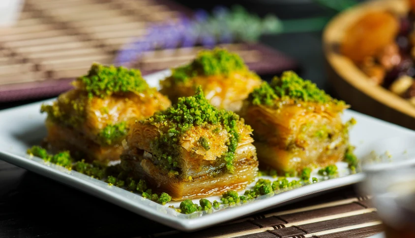 a close up of a plate of food on a table, hurufiyya, green and gold, pastry, square, hayao miyazak