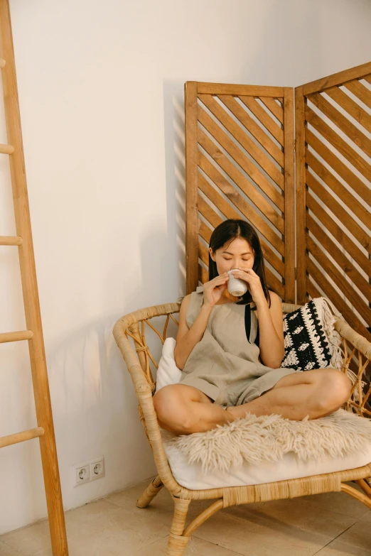 a woman sitting in a chair drinking from a cup, inspired by Li Di, pexels contest winner, tan skin a tee shirt and shorts, cozy calm! fabrics textiles, asian, wearing simple robes