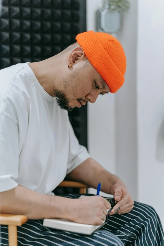 a man sitting in a chair writing on a piece of paper, by artist, pexels contest winner, with rap cap on head, studio orange, at checkout, book portrait