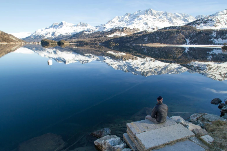 a person sitting on a rock next to a body of water, by Werner Andermatt, winter lake setting, water mirrored water, **cinematic, conde nast traveler photo