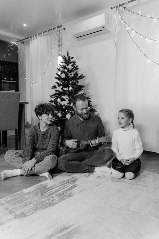 a black and white photo of a family sitting in front of a christmas tree, a black and white photo, pexels, figuration libre, thom yorke (guitar), indoor picture, threes, winter