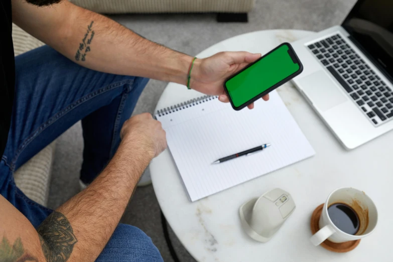 a man sitting at a table with a laptop and a cell phone, trending on pexels, happening, green and black, drawing on a parchment, no - text no - logo, iphone in hand