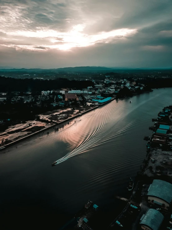 a boat traveling down a river under a cloudy sky, pexels contest winner, drone view of a city, laos, moody sunset, low quality footage