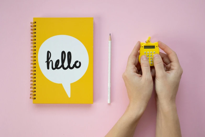 a person holding a calculator next to a notebook with the word hello written on it, a photo, by Paul Bird, art & language, pink and yellow, vinyl designer toy, hook as ring, official product photo