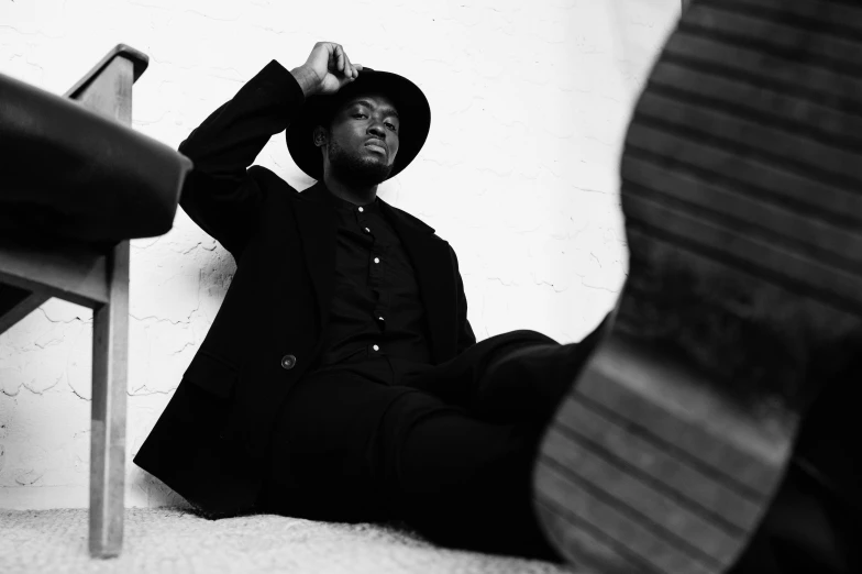a man sitting on the ground next to a chair, by Clifford Ross, pexels, black pointed hat, sharp black skin, doing an elegant pose, steven wiltshire