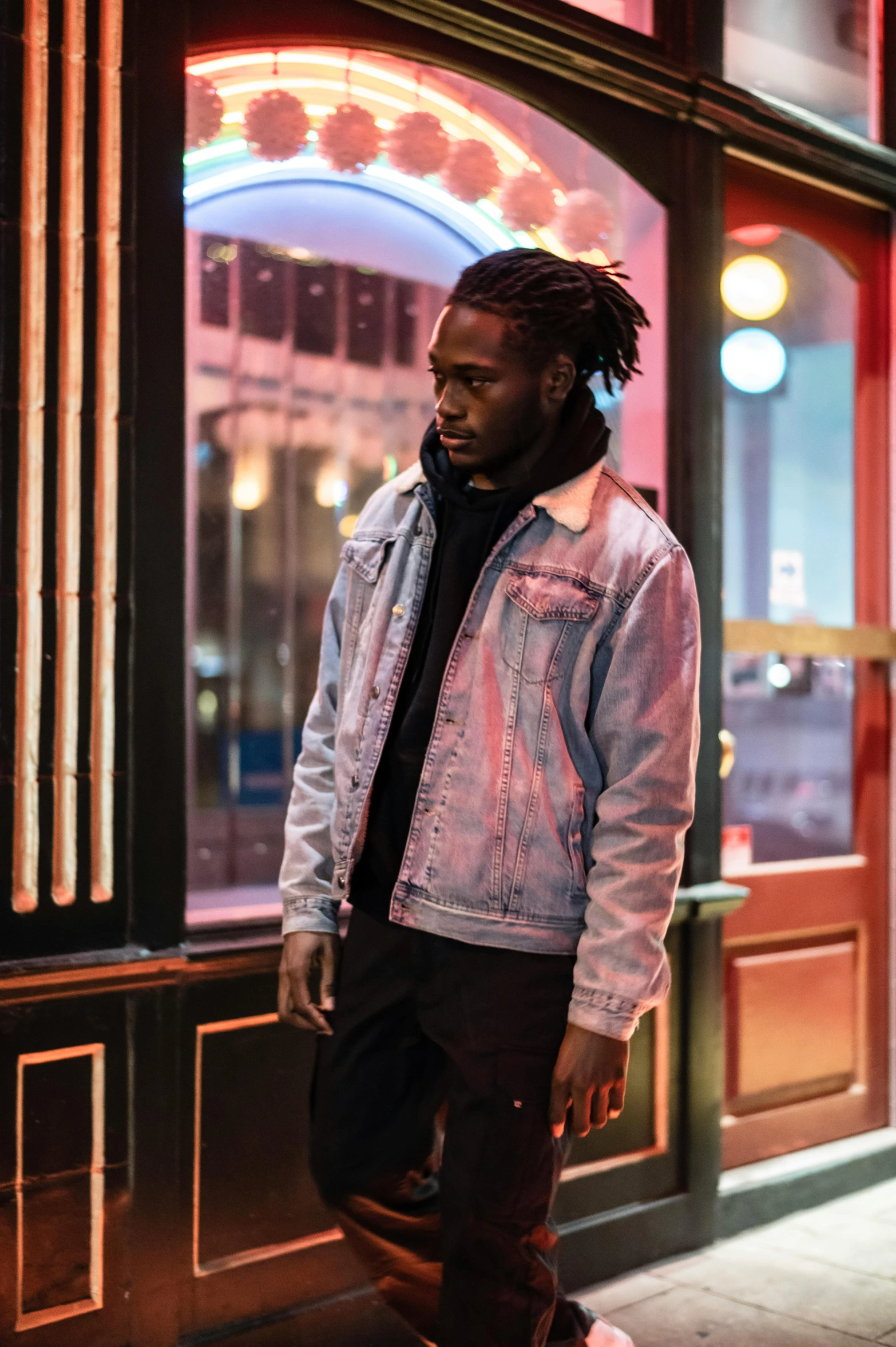 a man with dreadlocks standing in front of a building, an album cover, trending on pexels, visual art, wearing a turtleneck and jacket, night lights, jean jacket, ( ( dark skin ) )