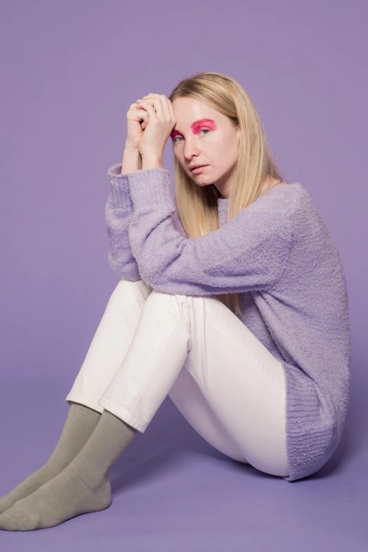 a woman sitting on the ground with her hand on her head, an album cover, inspired by Maud Naftel, trending on pexels, aestheticism, purple outfit, white pants, wearing casual sweater, color studio portrait